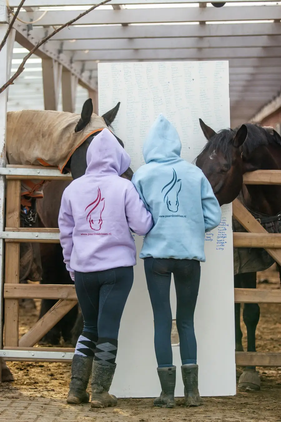 Twee dames in Paard Zoekt Baas-sweaters staan bij een deur waarop de namen staan van alle paarden die dankzij Paard Zoekt Baas een nieuw thuis hebben gevonden.
